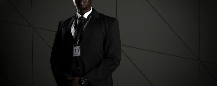 A person in a dark suit, wearing sunglasses and a security badge, stands against a geometric-patterned background. The lighting is low, creating a moody atmosphere.